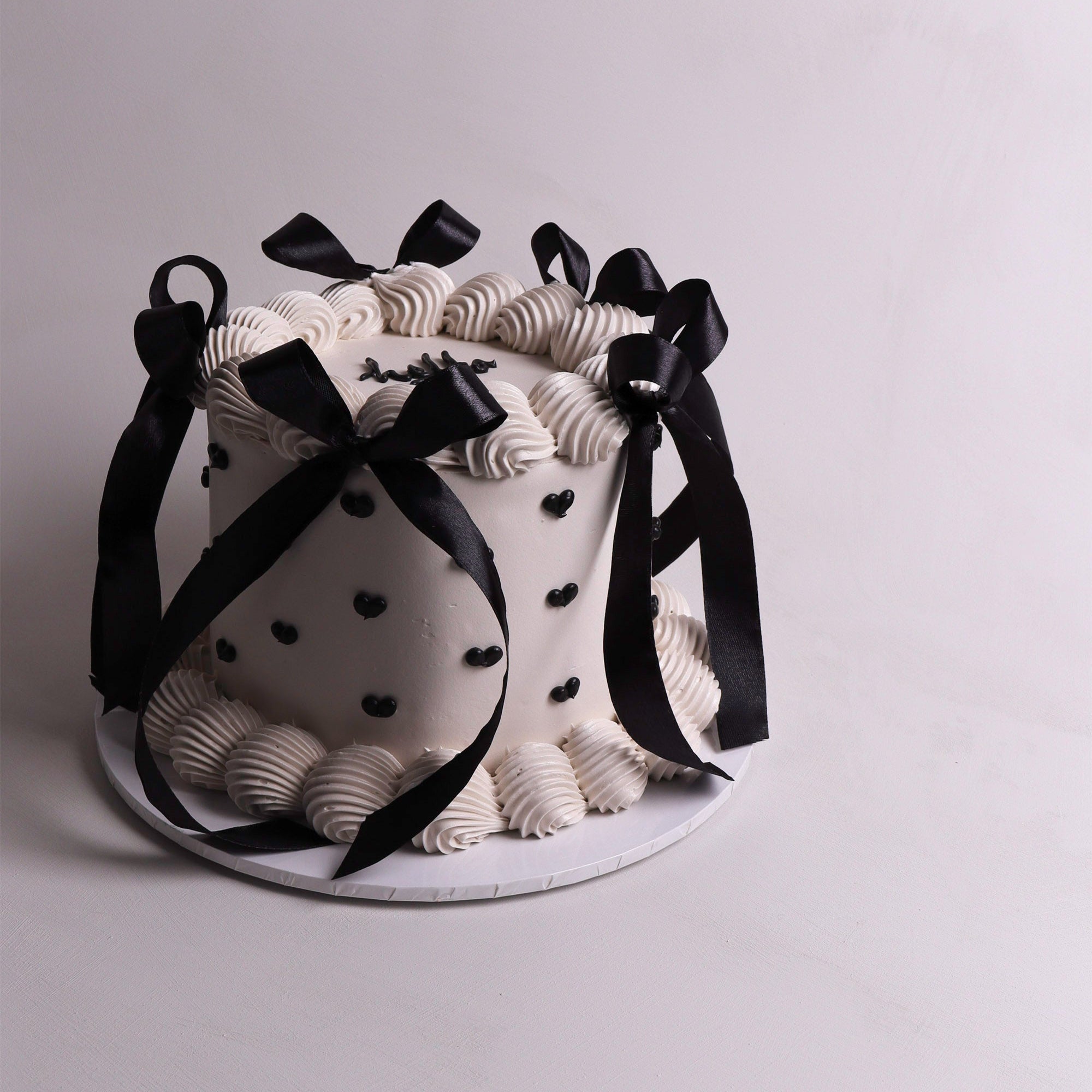 Round retro buttercream cake decorated with black satin ribbon bows, piped swirls and black heart accents – chic monochrome design for birthdays, parties and special events