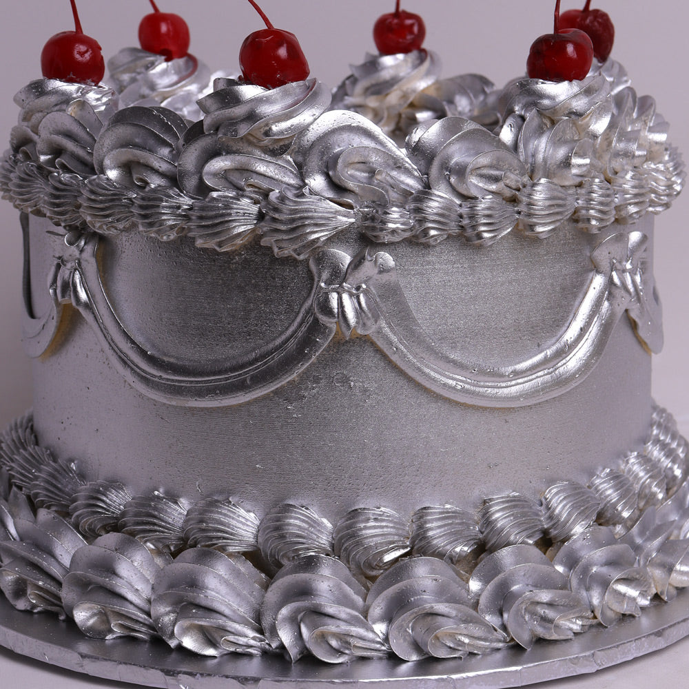 A silver cake decorated with intricate piped frosting patterns, topped with red cherries, shown on a silver cake board against a light background.