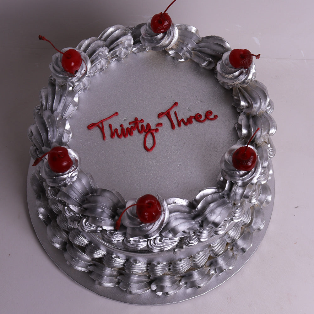 A round silver birthday cake decorated with piped frosting rosettes, red cherries on top, and red script text reading 'Thirty Three' on the top surface, photographed on a plain light background.