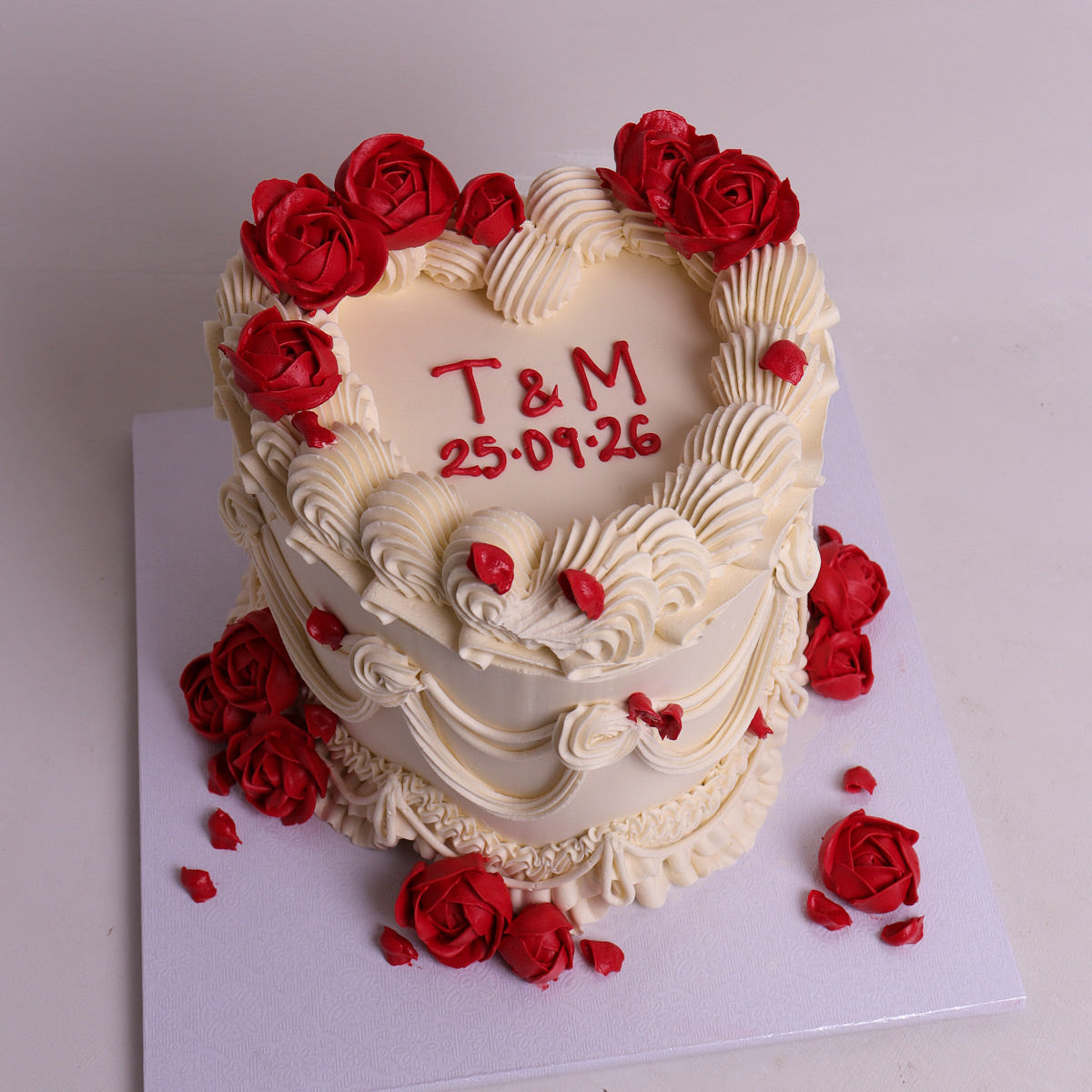 A heart-shaped white vintage style frosted cake decorated with red rose flowers and red icing details, with custom text written in red on the top, displayed on a white board against a light gray background.