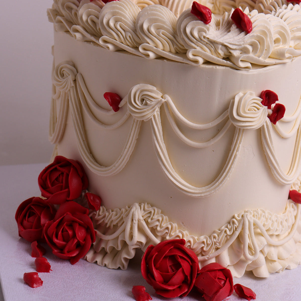 A romantic vintage cake with white piped frosting, draped scroll patterns, and red rose decorations on top and around the base, photographed against a plain light background.